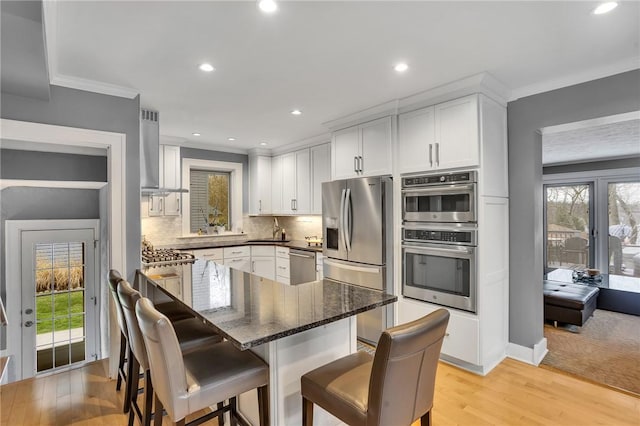 kitchen featuring ornamental molding, a breakfast bar area, wall chimney range hood, white cabinets, and appliances with stainless steel finishes