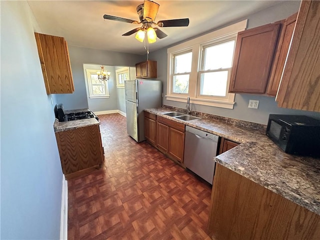 kitchen featuring dark countertops, appliances with stainless steel finishes, a sink, brown cabinetry, and ceiling fan with notable chandelier