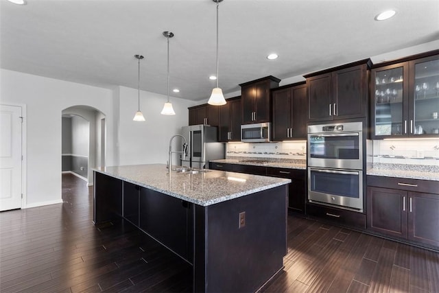 kitchen with light stone countertops, an island with sink, dark wood-type flooring, arched walkways, and appliances with stainless steel finishes