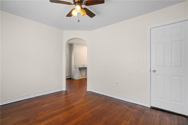 empty room with baseboard heating, arched walkways, a ceiling fan, baseboards, and wood finished floors
