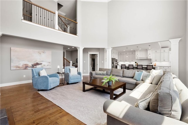 living room featuring stairs, decorative columns, a towering ceiling, wood finished floors, and baseboards