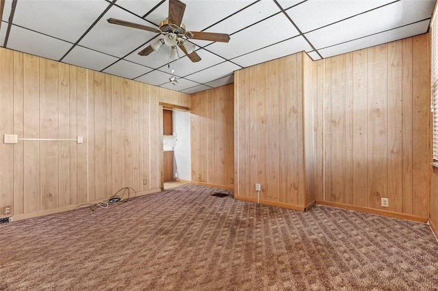 empty room featuring a ceiling fan, a paneled ceiling, baseboards, wooden walls, and carpet flooring