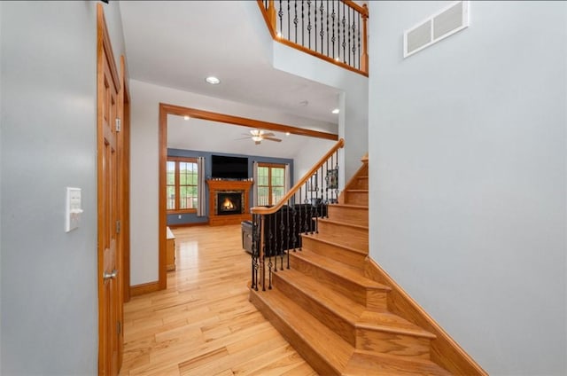 stairway with a warm lit fireplace, a ceiling fan, baseboards, wood finished floors, and visible vents