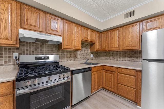 kitchen with a sink, ornamental molding, under cabinet range hood, light countertops, and appliances with stainless steel finishes