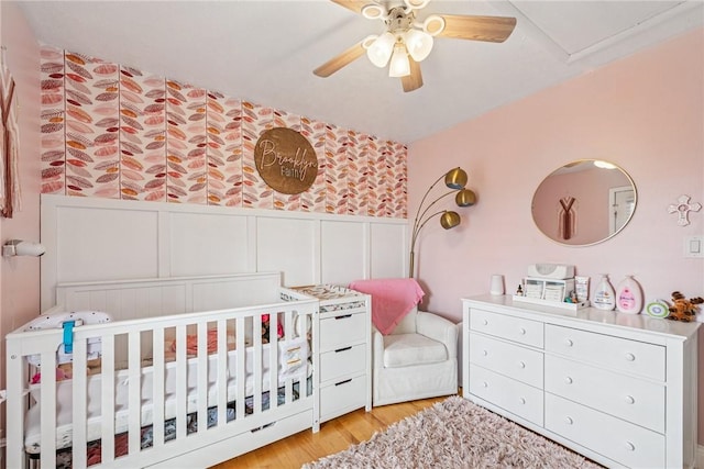 bedroom with a ceiling fan, a wainscoted wall, a crib, light wood-type flooring, and a decorative wall