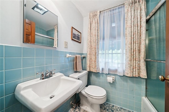 bathroom featuring tile walls, toilet, bath / shower combo with glass door, a sink, and a wainscoted wall