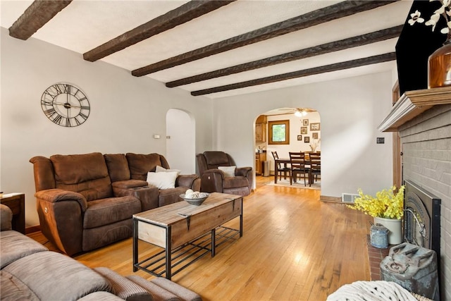 living area featuring beam ceiling, visible vents, a brick fireplace, arched walkways, and light wood-style floors