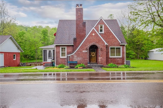 tudor-style house featuring a front lawn, a shingled roof, brick siding, and a chimney