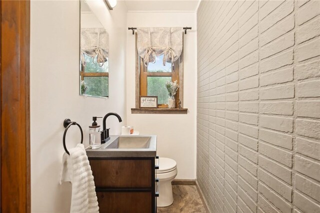 bathroom with toilet, brick wall, baseboards, an accent wall, and vanity
