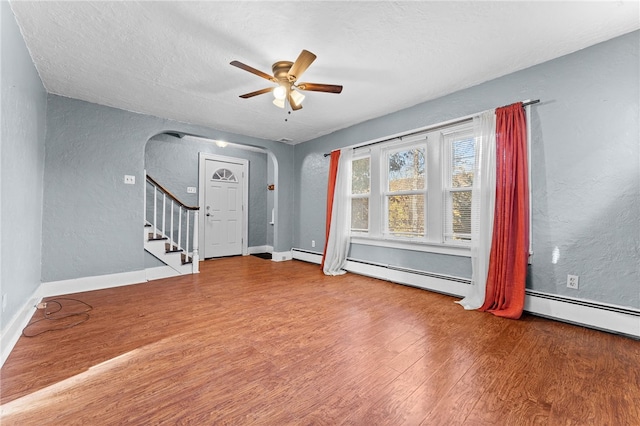 empty room featuring arched walkways, wood finished floors, a textured wall, a ceiling fan, and a baseboard radiator