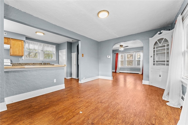 unfurnished living room featuring a ceiling fan, wood finished floors, arched walkways, baseboards, and a baseboard heating unit