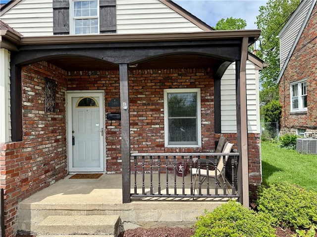 view of exterior entry featuring central AC unit, brick siding, covered porch, and fence