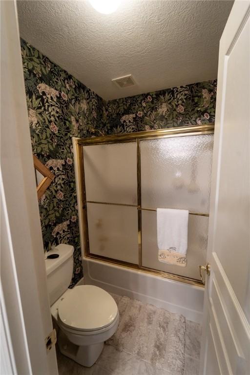 full bath with wallpapered walls, a textured ceiling, shower / bath combination with glass door, and toilet