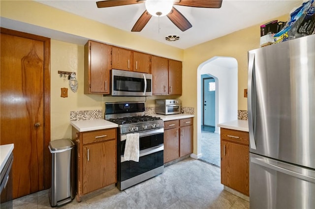 kitchen featuring stainless steel appliances, brown cabinetry, light countertops, ceiling fan, and arched walkways