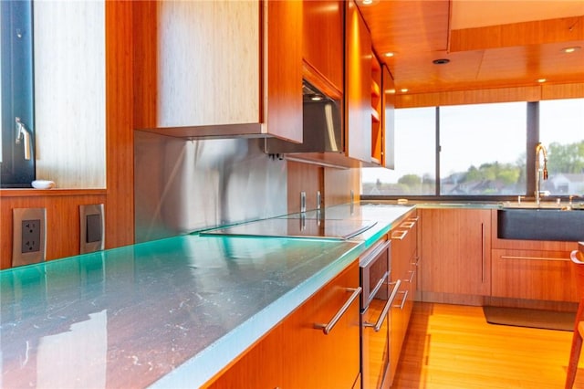 kitchen featuring a sink, black electric cooktop, oven, light wood finished floors, and brown cabinetry