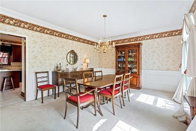 dining room with a chandelier, wainscoting, wallpapered walls, light colored carpet, and crown molding