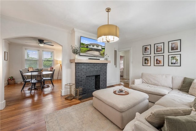 living room featuring arched walkways, crown molding, wood finished floors, a ceiling fan, and a brick fireplace