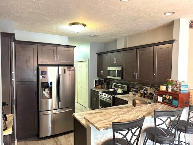 kitchen with appliances with stainless steel finishes, a sink, dark brown cabinets, a breakfast bar, and a textured ceiling
