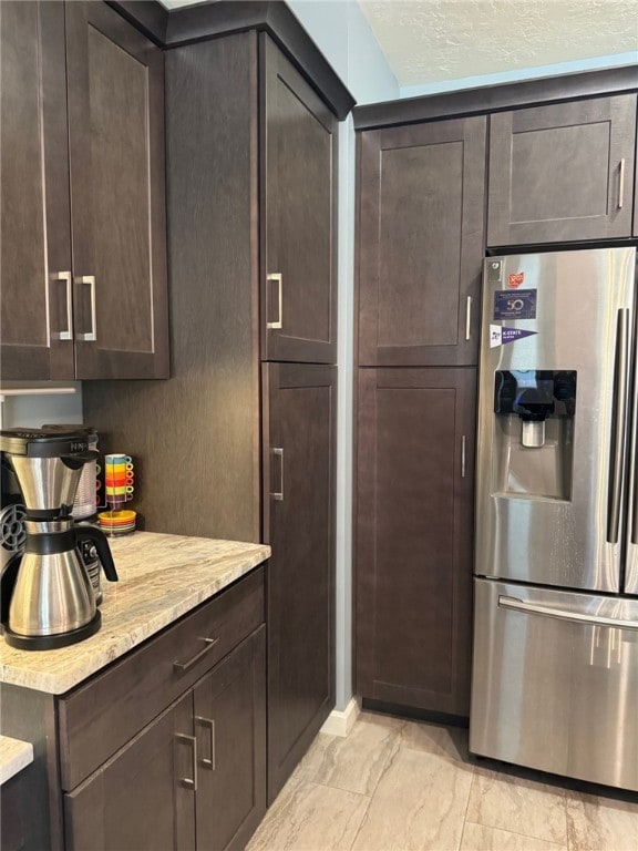 kitchen featuring stainless steel fridge, dark brown cabinets, light marble finish floors, light stone countertops, and a textured ceiling