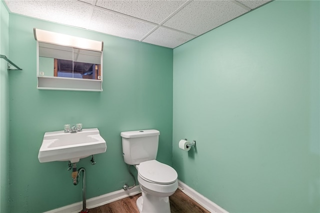 half bath featuring wood finished floors, toilet, a drop ceiling, a sink, and baseboards