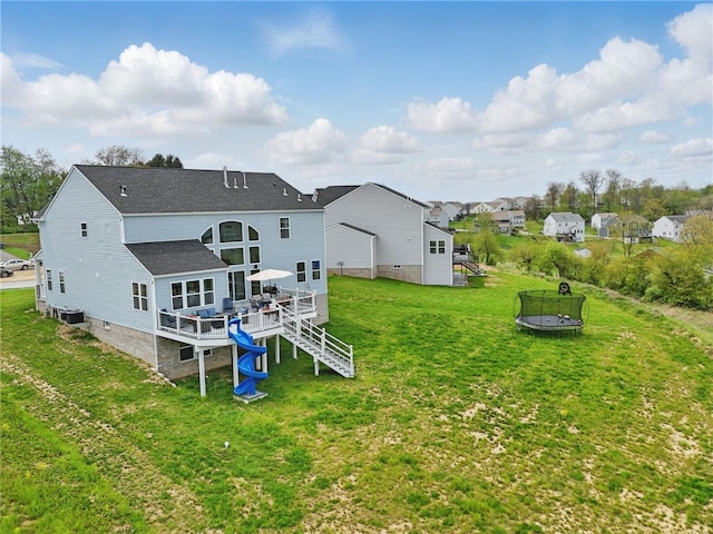 back of property with a trampoline, a deck, central air condition unit, a yard, and roof with shingles