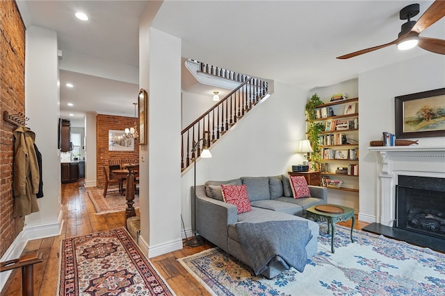 living room featuring wood-type flooring, a fireplace with raised hearth, stairs, a ceiling fan, and brick wall