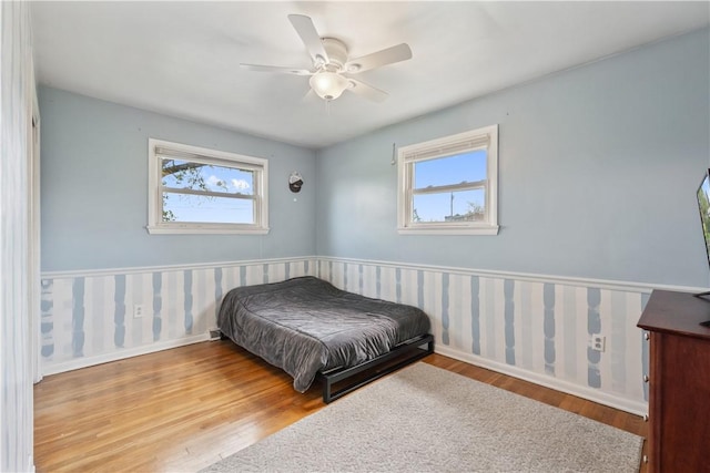 bedroom with wainscoting, wood finished floors, wallpapered walls, baseboards, and a ceiling fan