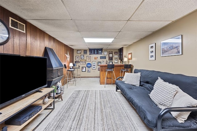 living room featuring a dry bar, a drop ceiling, wooden walls, and light colored carpet