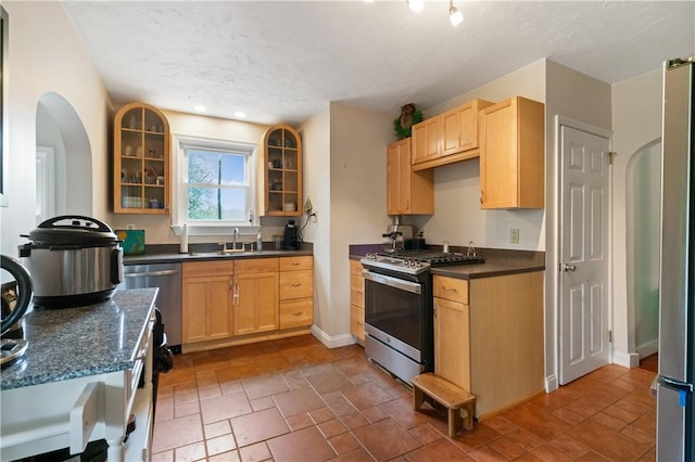 kitchen featuring appliances with stainless steel finishes, light brown cabinetry, dark countertops, glass insert cabinets, and a sink