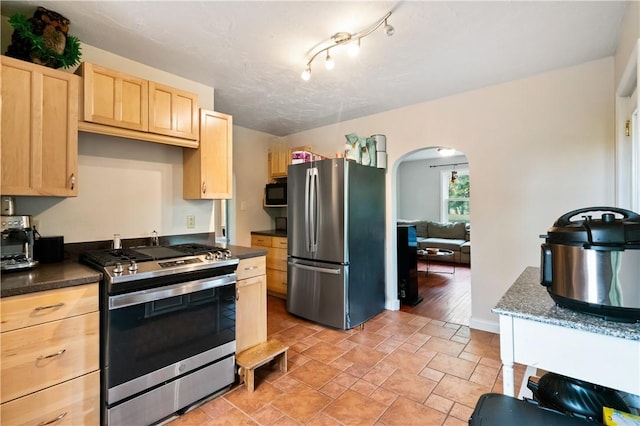 kitchen featuring stainless steel appliances, light brown cabinetry, arched walkways, and dark countertops