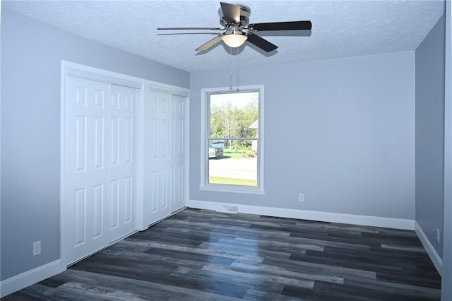 unfurnished bedroom with dark wood-type flooring, two closets, a textured ceiling, baseboards, and a ceiling fan