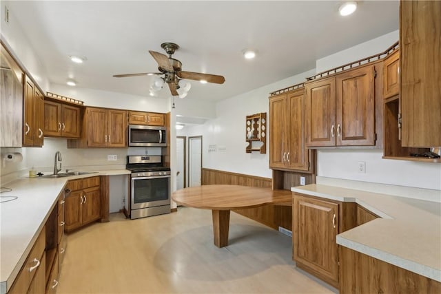 kitchen featuring stainless steel appliances, a sink, a wainscoted wall, ceiling fan, and brown cabinetry