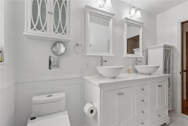 bathroom with wainscoting, double vanity, toilet, and a decorative wall