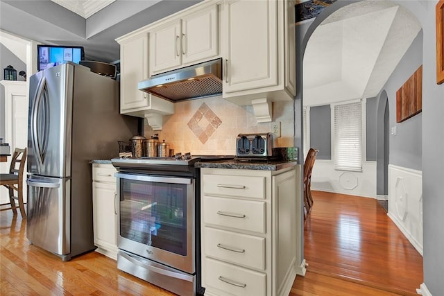 kitchen featuring appliances with stainless steel finishes, under cabinet range hood, light wood finished floors, arched walkways, and backsplash