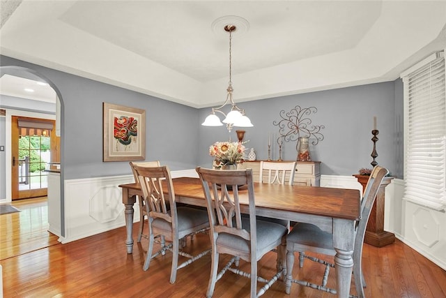 dining space with a raised ceiling, arched walkways, wood finished floors, and a wainscoted wall