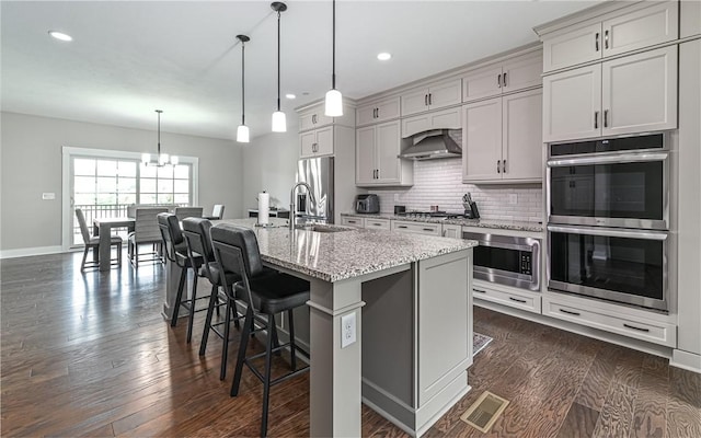 kitchen with appliances with stainless steel finishes, wall chimney range hood, a chandelier, a center island with sink, and a breakfast bar area