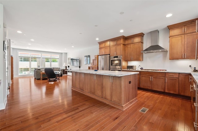 kitchen with wall chimney exhaust hood, stainless steel appliances, brown cabinetry, open floor plan, and a center island