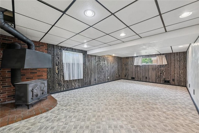 below grade area featuring a wood stove, baseboards, wood walls, carpet floors, and a drop ceiling