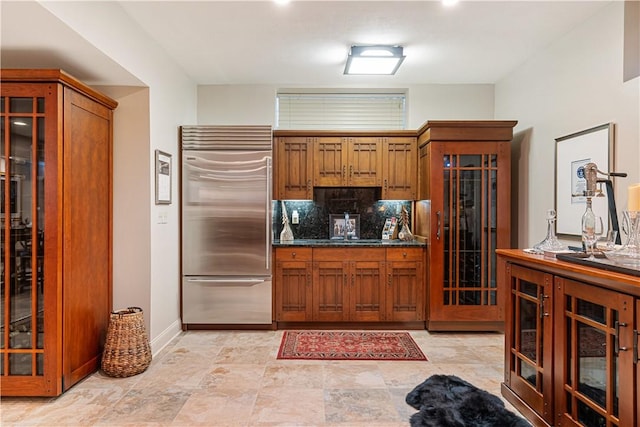 kitchen with stainless steel built in refrigerator, dark countertops, brown cabinets, baseboards, and glass insert cabinets