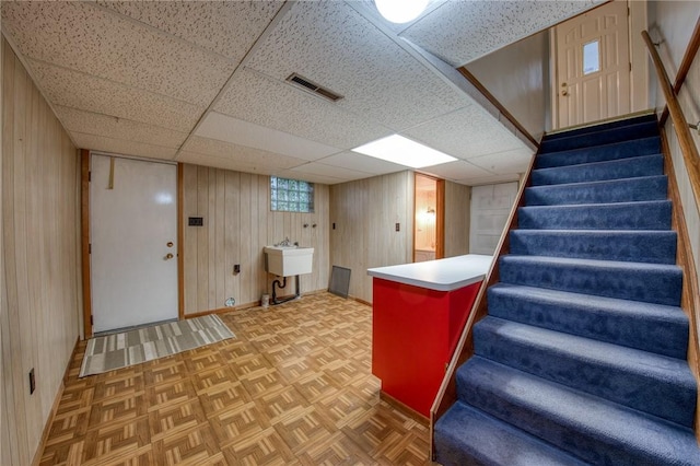 below grade area with wooden walls, stairs, baseboards, a sink, and a drop ceiling