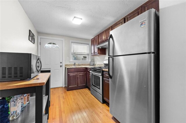 kitchen with stainless steel appliances, under cabinet range hood, butcher block countertops, light wood-type flooring, and a textured ceiling