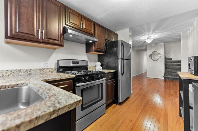 kitchen with appliances with stainless steel finishes, under cabinet range hood, light wood finished floors, dark brown cabinets, and light stone countertops