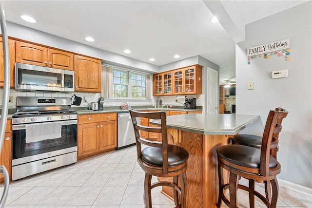 kitchen with appliances with stainless steel finishes, a breakfast bar area, glass insert cabinets, brown cabinets, and recessed lighting