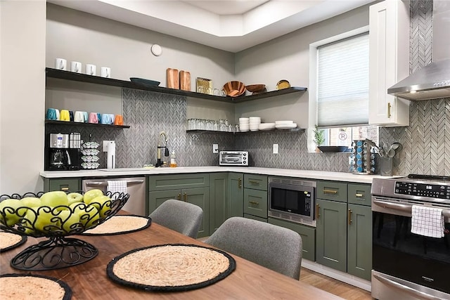 kitchen featuring stainless steel appliances, wall chimney range hood, green cabinets, open shelves, and light countertops