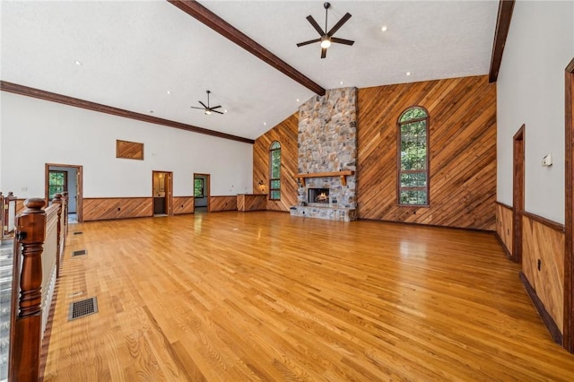 unfurnished living room featuring ceiling fan, wooden walls, beamed ceiling, a stone fireplace, and light wood-style flooring