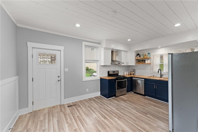 kitchen with stainless steel appliances, wall chimney range hood, butcher block countertops, blue cabinetry, and a sink