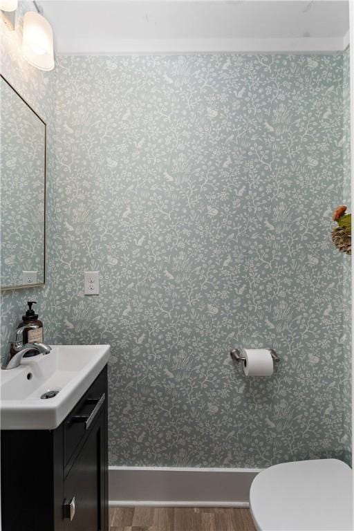 half bathroom with wood finished floors, vanity, toilet, wallpapered walls, and baseboards