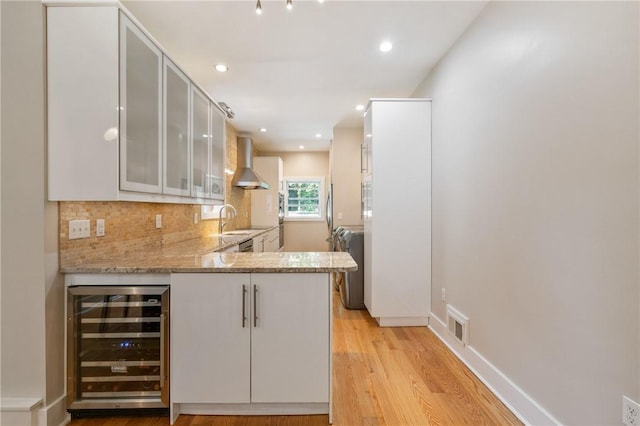 kitchen with wine cooler, wall chimney exhaust hood, light stone countertops, backsplash, and light wood-style floors
