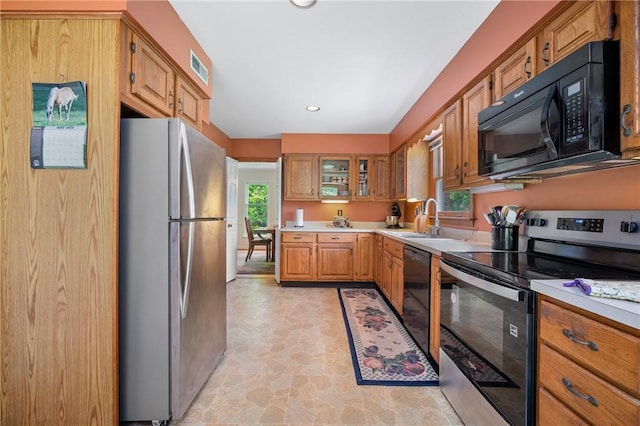 kitchen with black appliances, a sink, light countertops, brown cabinetry, and glass insert cabinets