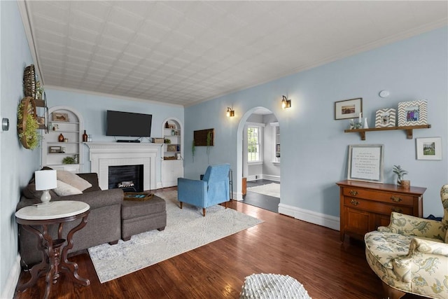 living room featuring arched walkways, a brick fireplace, wood finished floors, baseboards, and ornamental molding
